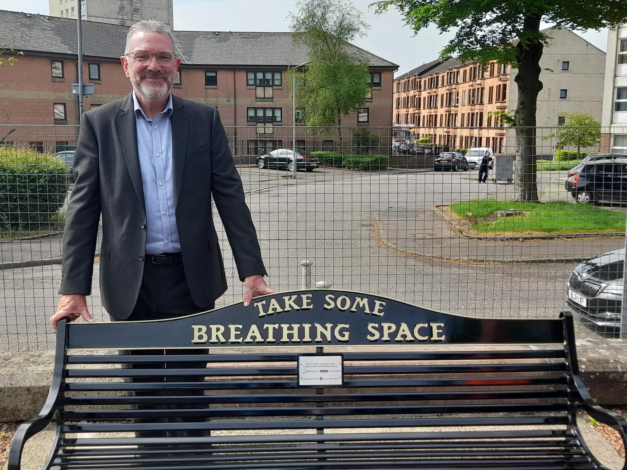 man standing behind iron bench on train station platform. Bench is black with gold lettering that says take some breathing space in gold.